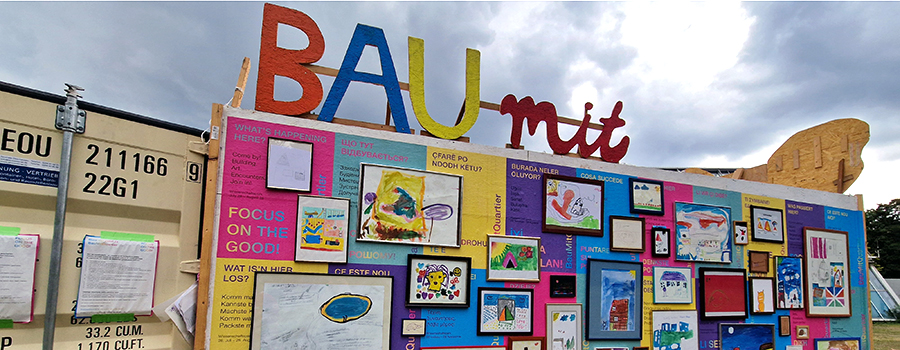 Bunter Baucontainer mit Holzbuchstaben "Bau mit" und einer Bildergalerie, mit Werken von Kindern